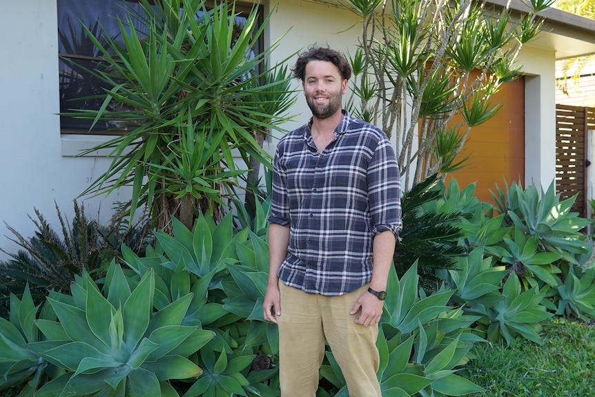 A man in beige chinos and a navy and white checked shirt stands in front of a garden.