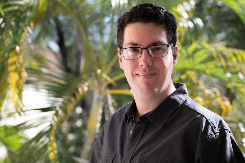 Mr Nicholls is pictured with palm trees behind him. He's wearing glasses and a back shirt.