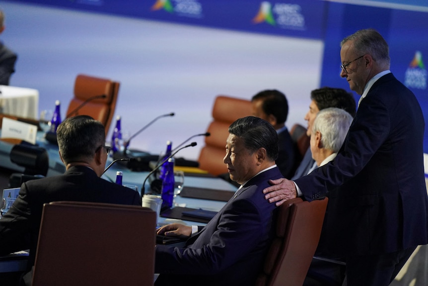 Anthony Albanese stands while putting a hand on the shoulder of Xi Jinping, who is sitting at a round desk
