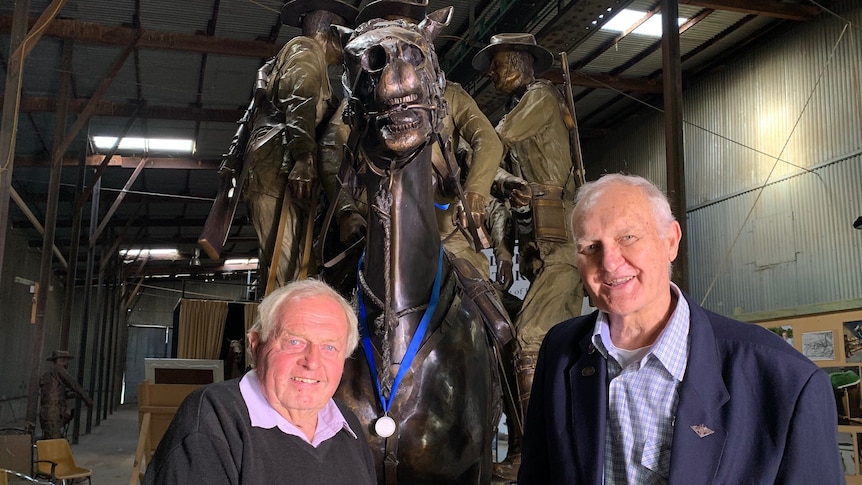 Two elderly men with grey hair stand in front of a bronze horse statue and smile for photo. 