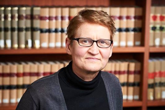 A woman with auburn hair and glasses standing with a bookshelf in the background.