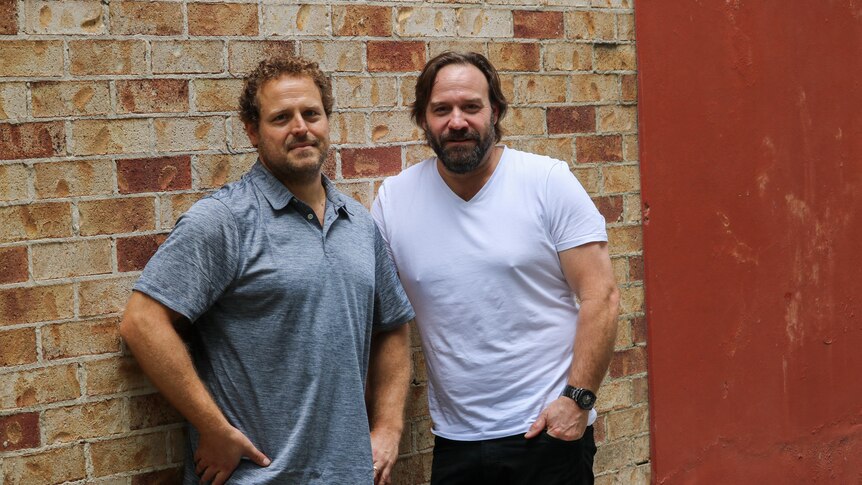 Joel Spreadborough and John Manning standing next to each other in front of a brick wall.