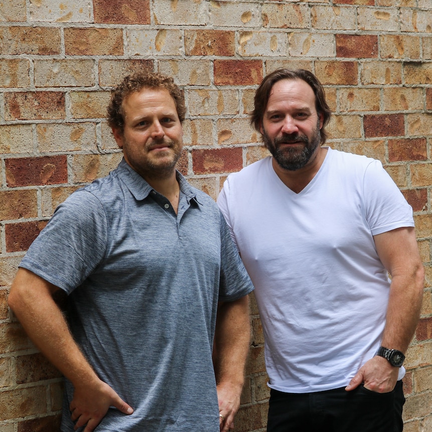 Joel Spreadborough and John Manning standing next to each other in front of a brick wall.