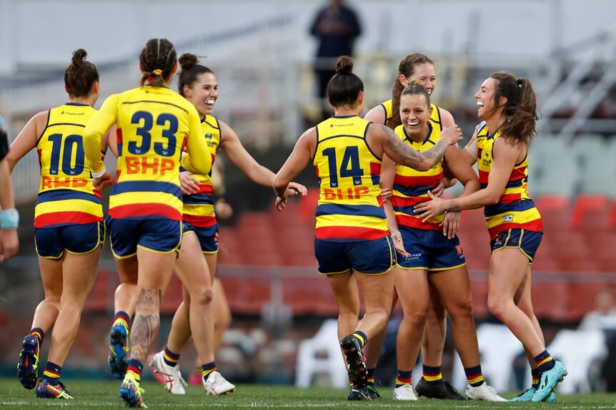A group of Adelaide Crows AFLW players converge on a smiling teammate who has just kicked a goal.