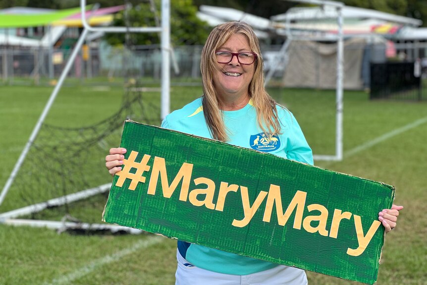 A woman with long hair wearing a turquoise soccer shirt holding a sign that says #MaryMary