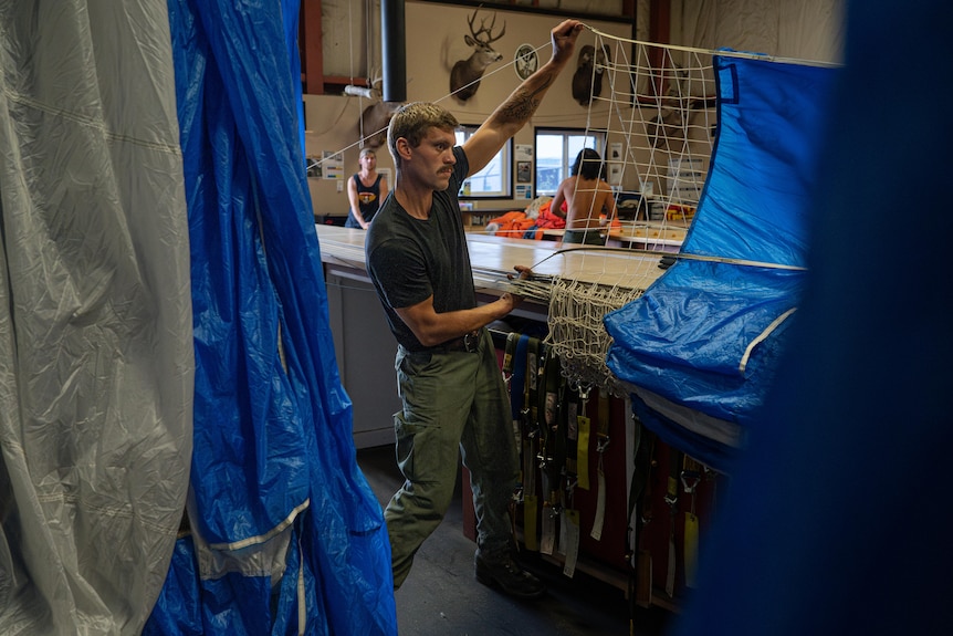 A man in a workshop holds up and stretches out the netting of a parachute, checking it's not tangled.