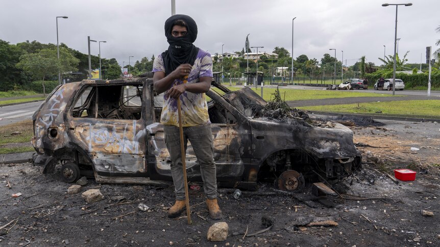 A man wearing a black cloth to cover most of his face stands leaning on a stick in front of a burnt-out car