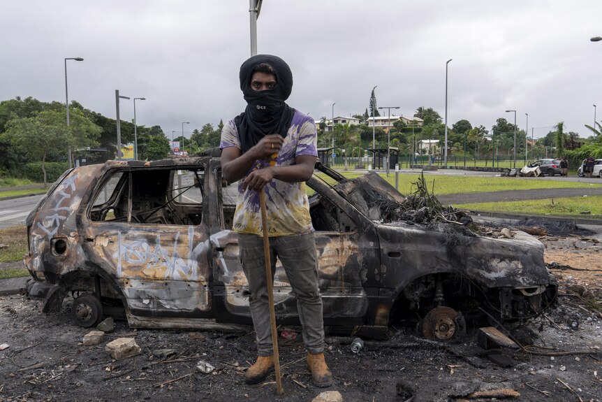 A man wearing a black cloth to cover most of his face stands leaning on a stick in front of a burnt-out car