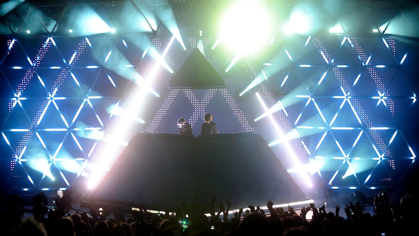 Two robots atop a massive pyramid structure DJ to a crowd. There are lots of flashing lights around them.