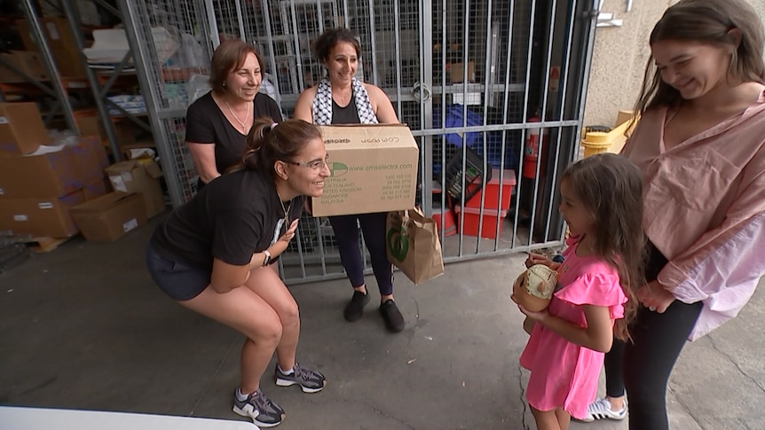 A little girl in a pink dress is seen giving her piggy bank to volunteers in a warehouse.