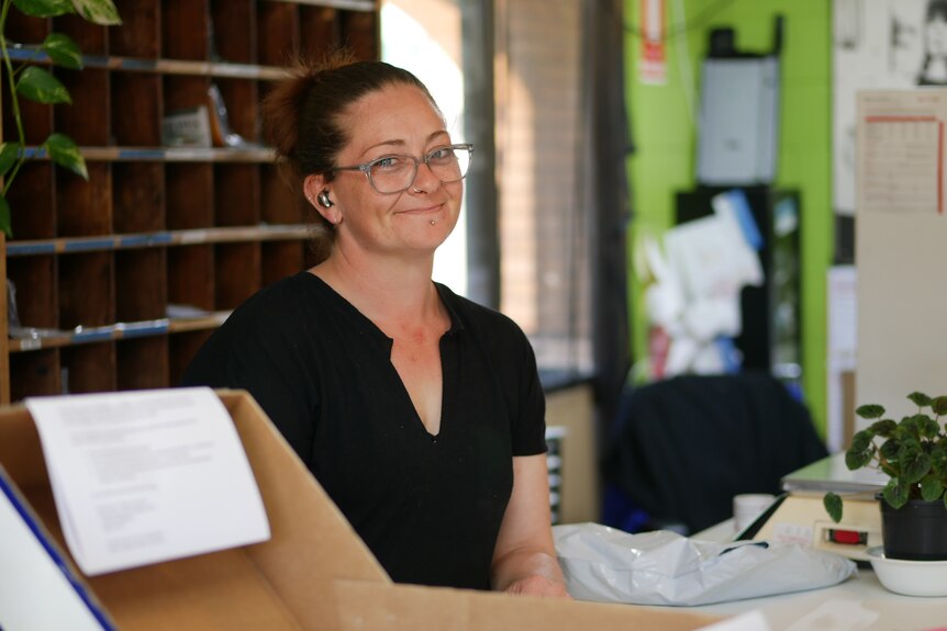 A woman with glasses and wearing ear buds smiles behind a post office counter. 