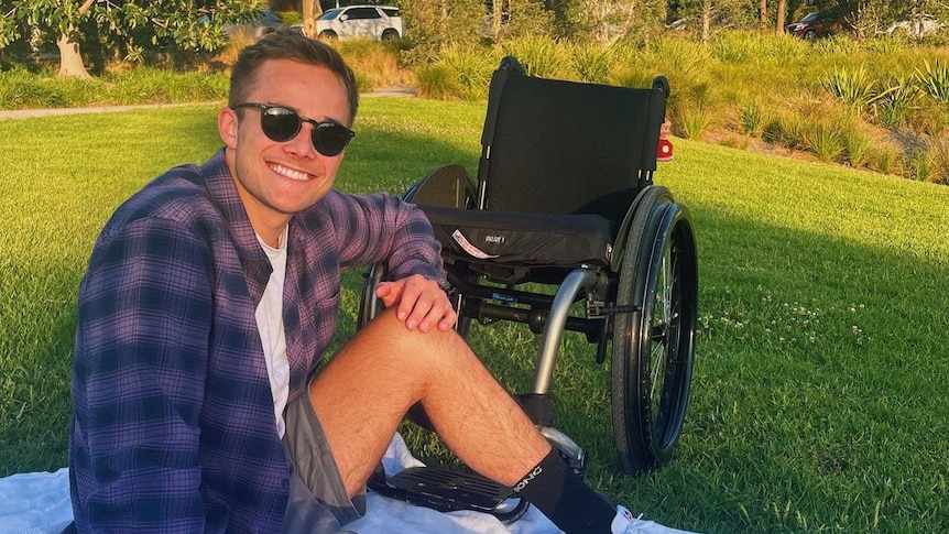 A man pictured smiling on a picnic blanket in a park next to a wheelchair.