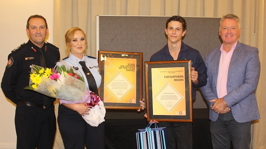 Two award recipients holding up their framed awards flanked by dignitaries at an awards ceremony.