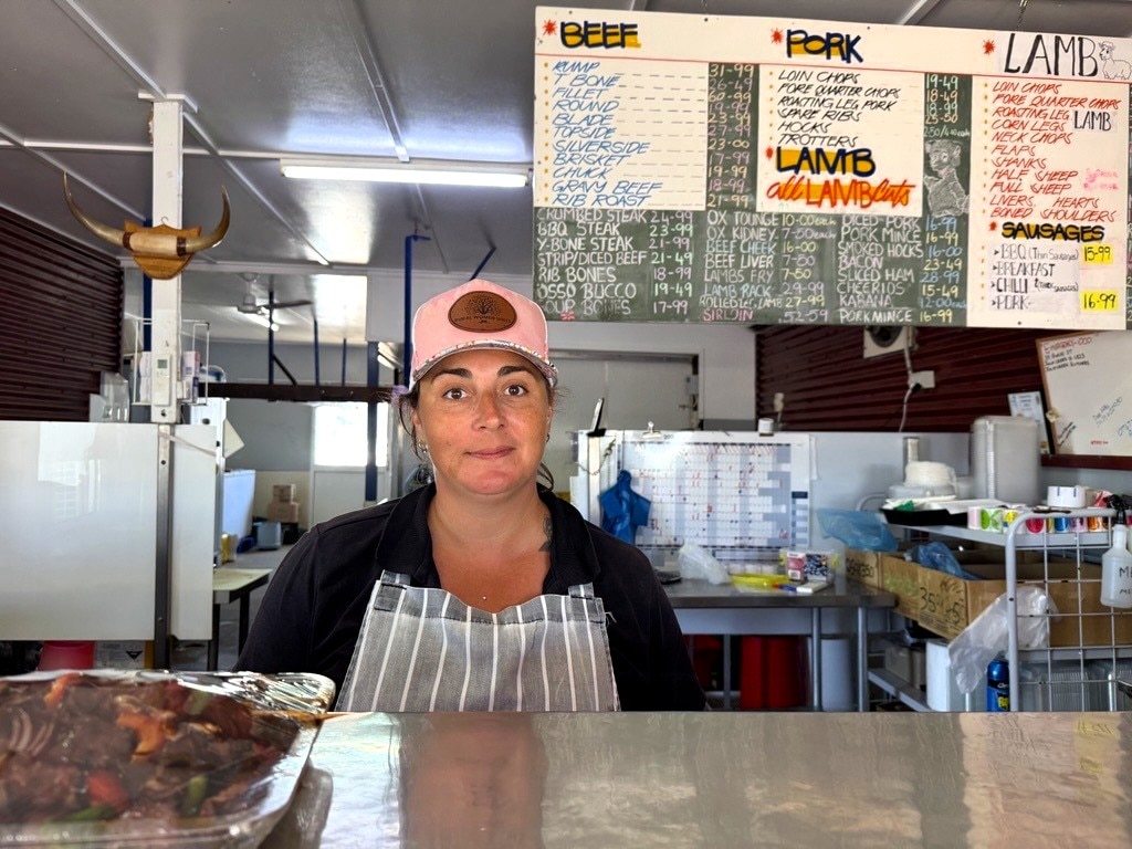 Female butcher in cap and apron standing behind the counter of her store with kebabs in front of her and beef/lamb sign.