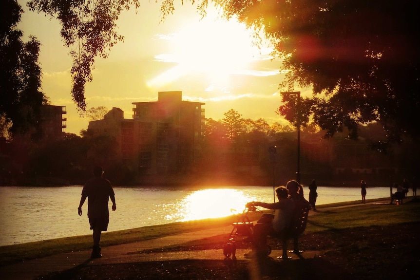 Sun sets over Brisbane river with people sitting on a bench and a man walking.