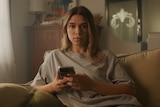 A young woman sits on a couch looking at a mobile phone.