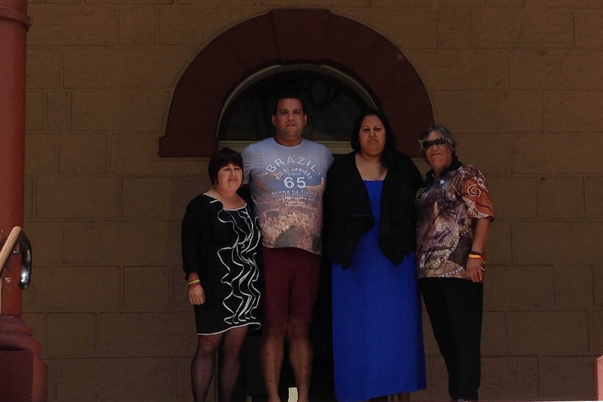 Four Indigenous people stand in front of a historic-looking building with their arms around each other.