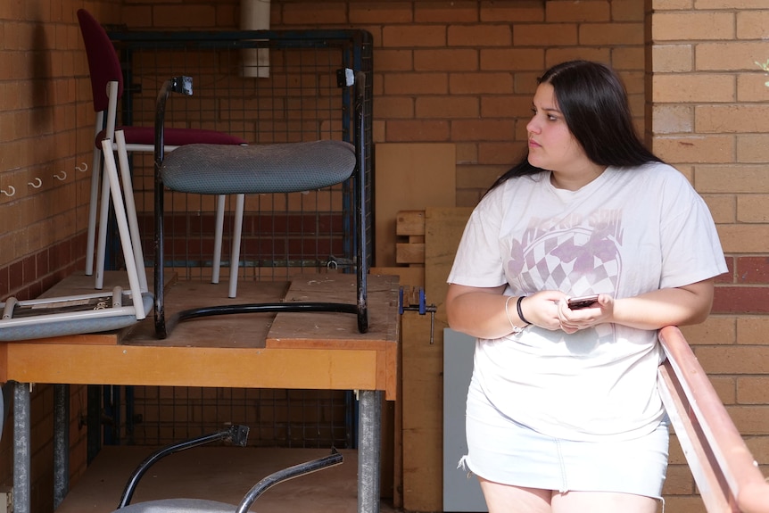 A teenage girl wearing a denim skirt and white t-shirt holds her phone and looks to her side.