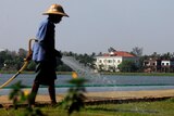 A picture of a woman watering with a hose with a large house in the background with a view of the Inya Lake