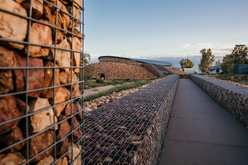 A building stands made out of red rocks stacked within wire enclosures.