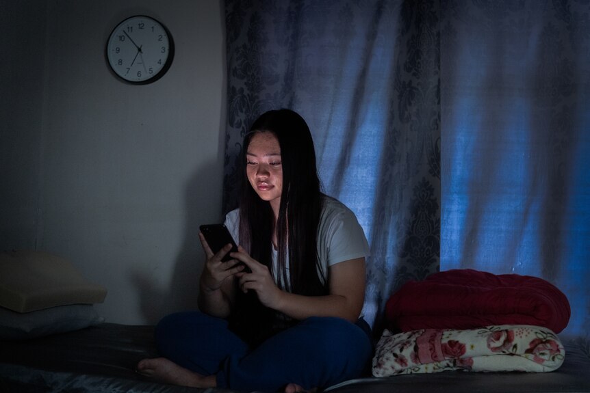 Teenage girl looks at phone in dark bedroom.