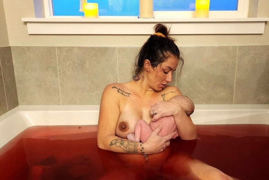 A woman sits in a bathtub full of bloody water holding her newborn baby