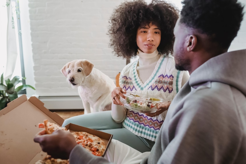 Man and woman sitting on couch eating pizza as the woman listens