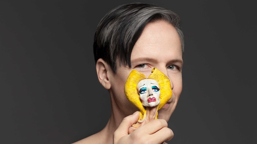 John Cameron Mitchell, in front of a grey backdrop, holds a small bust of his character Hedwig in front of his face.