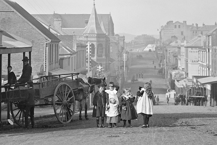 A vintage black and white photo of 5 children standing near a horse cart on the street, old buildings in the background