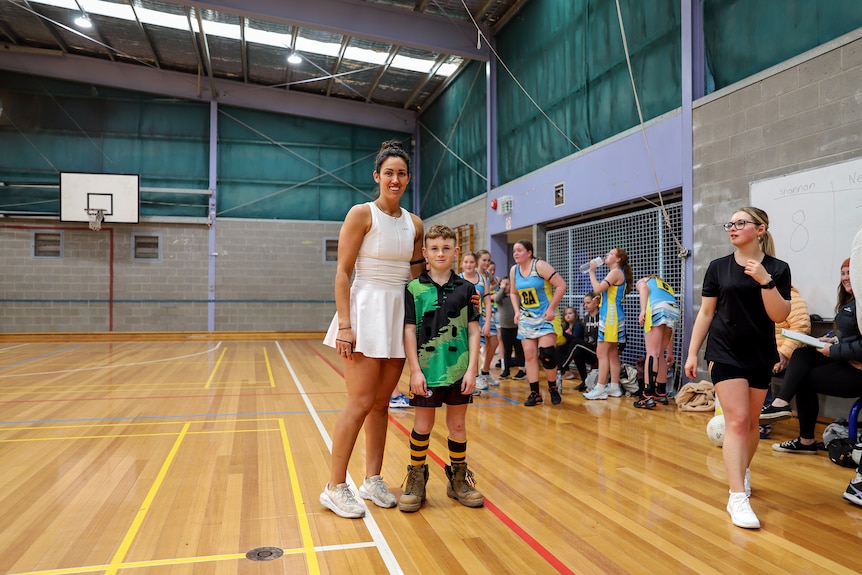 Woman wearing white netball referee outfit and her younger son wearing boots and a netball uniform on a indoor basketball court.
