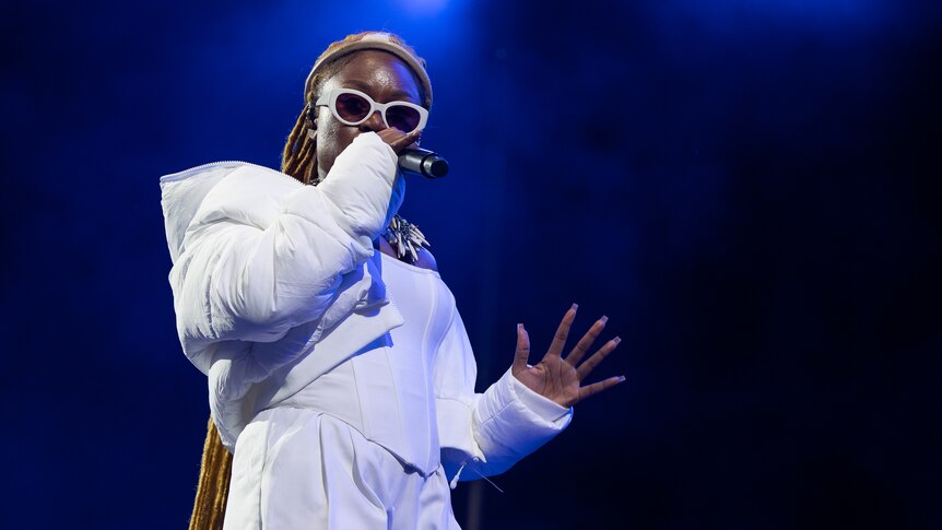 Sampa The Great raps into a microphone on stage wearing all white and sunglasses
