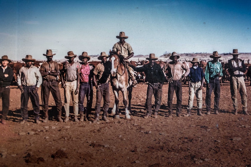 A group of stockmen stand in a line with one mounted on a horse.