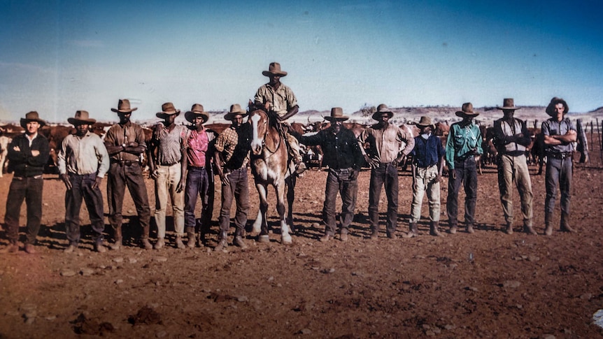 A group of stockmen stand in a line with one mounted on a horse.