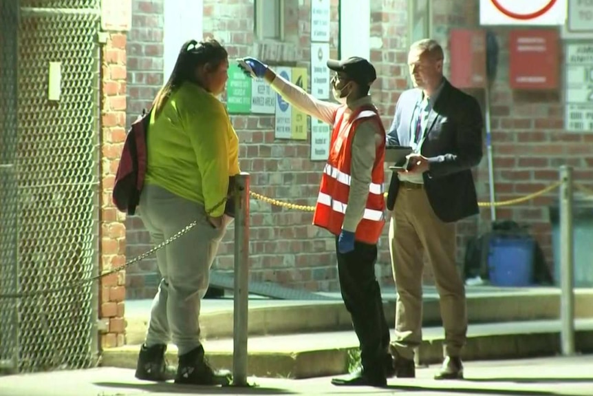 A woman stands outside a brick building getting her temperature tested by a man in a high-vis vest under fluorescent lights.