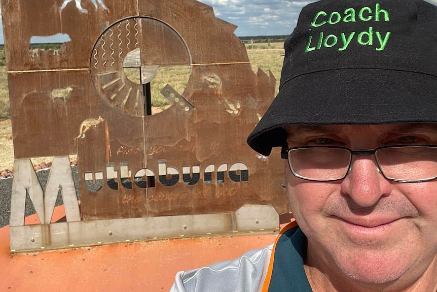 A bloke with glasses and a "Coach Lloydy" bucket hat stands in front of a rusty metal sculpture of Qld with the word Muttaburra 