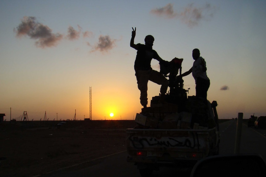 Libyan fighters stand on top of a combat vehicle as the sun sets behind them.