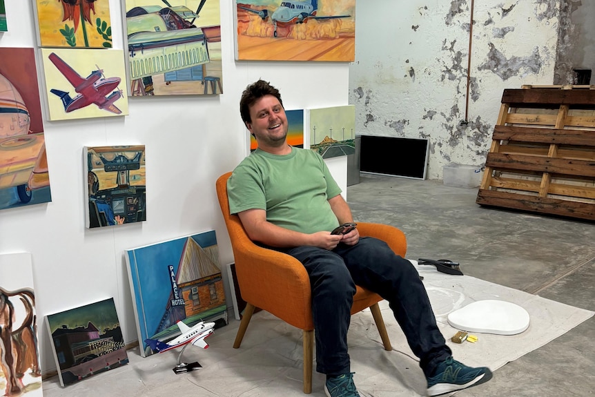 A man in a green shirt sits in an orange chair in front of a wall of paintings.