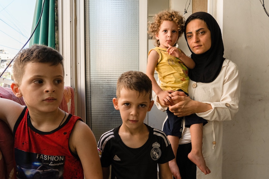 A woman wearing a black hijab with a white dress holds onto her baby as her children stand in front of her.