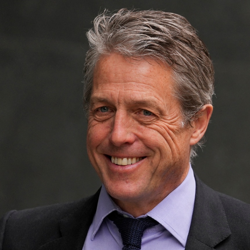 actor Hugh Grant smiles while wearing a blue button up shirt and a navy blue tie.