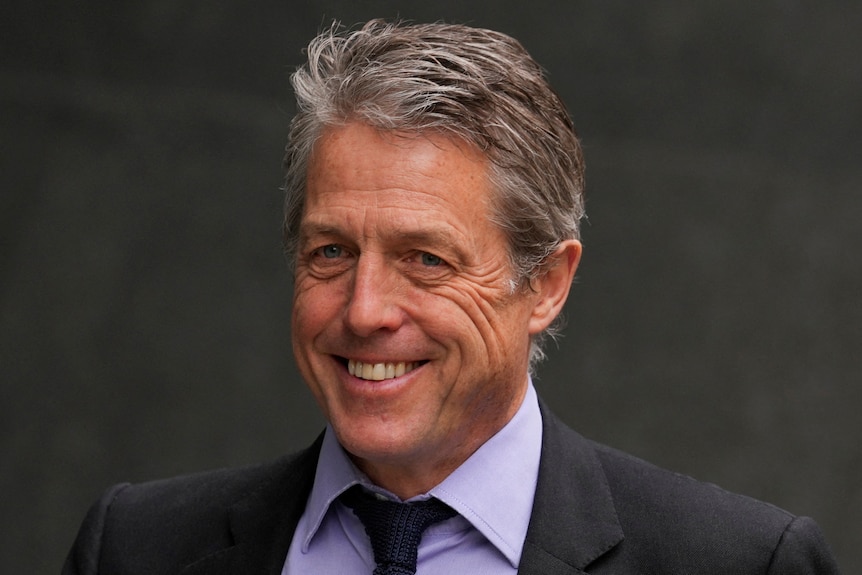 actor Hugh Grant smiles while wearing a blue button up shirt and a navy blue tie.
