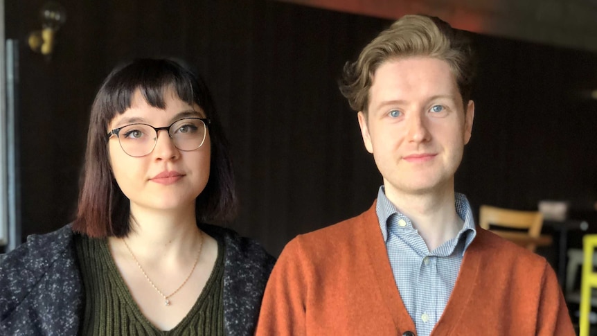 A young couple sit holding hands and looking into the camera.