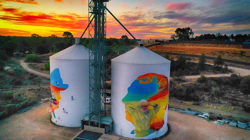 Two silos with colourful paintings are backdropped by a beautiful swirling sunset of red, orange, yellow, blue, pink and purple.