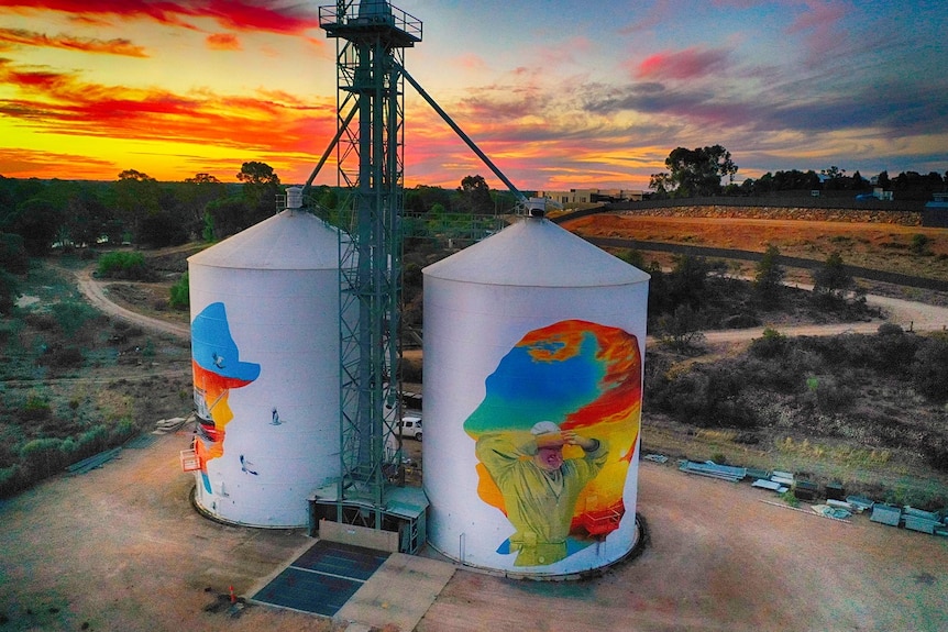 Two silos with colourful paintings are backdropped by a beautiful swirling sunset of red, orange, yellow, blue, pink and purple.