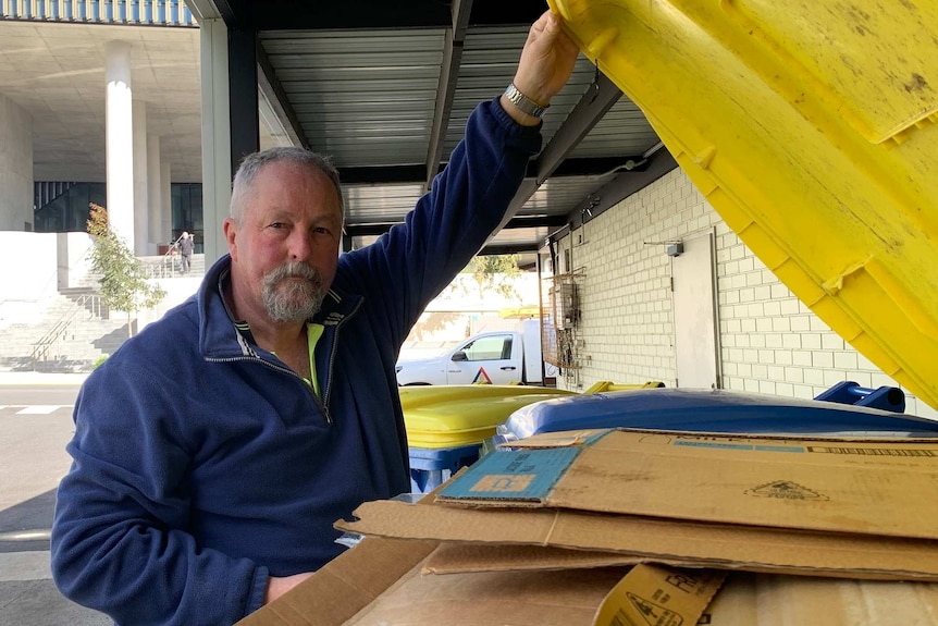 A man stands in front of yellow and blue recycling bins lifting the lid of one with his hand and looking at the camera