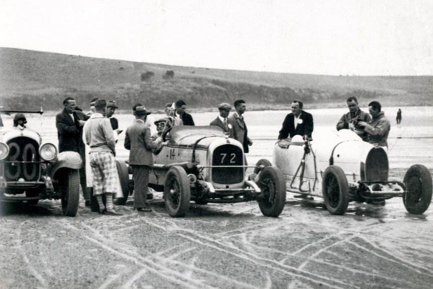 Men stand around admiring vintage cars on the beach