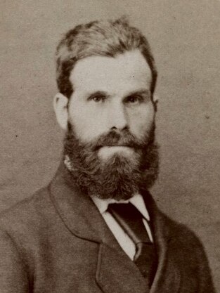 A historic, clack-and-white portrait photograph of a young bearded man in a suit.
