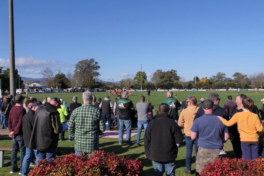 Local crowds stand around watching an Australian Rules football game at Whorouly.
