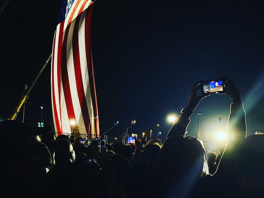 Night shot of people holding up phones filming Biden in distance and large US flag waving.