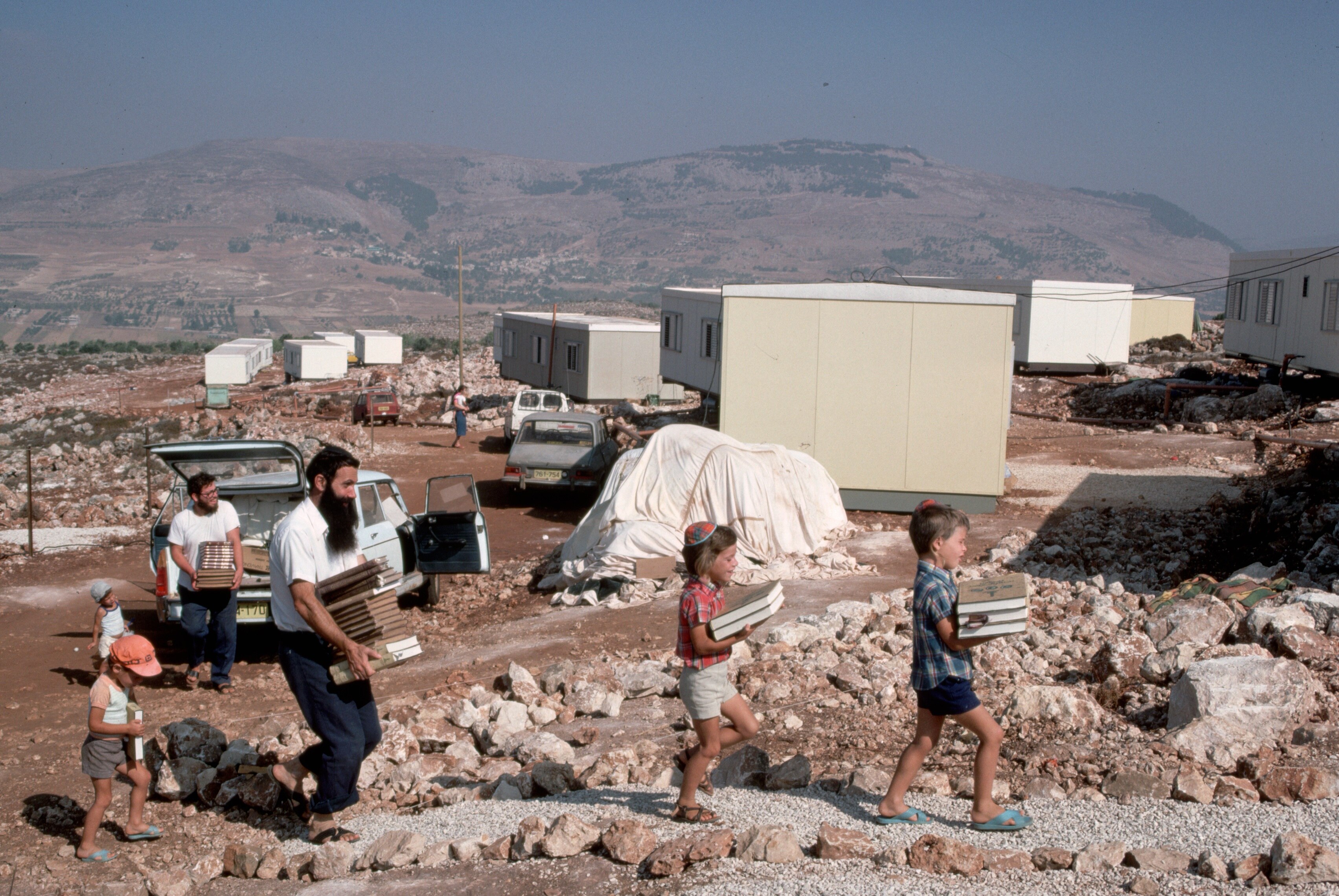 A family unloads a car and walks towards temporary housing, in the desert.