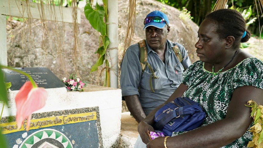 A man and a woman look solemnly at a tombstone